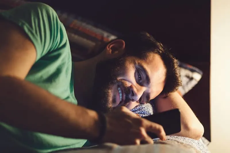 homme barbu heureux allongé sur le lit et regardant son téléphone